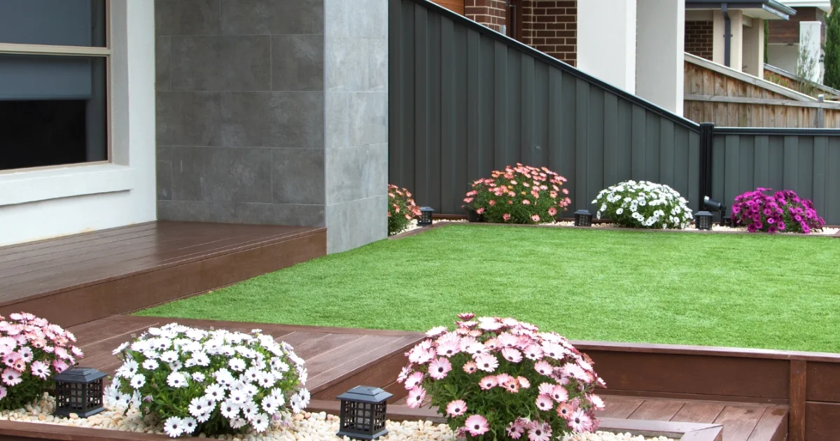 Well-maintained synthetic lawn with colorful flower beds and a modern wooden deck.